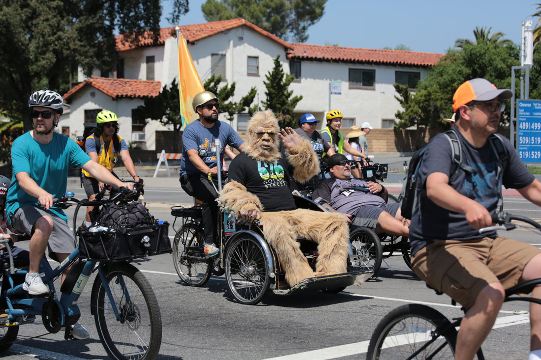 Photo of cyclists on the street. Gabe, a large sasquatch, is seated in the cab of a pedi-cab.